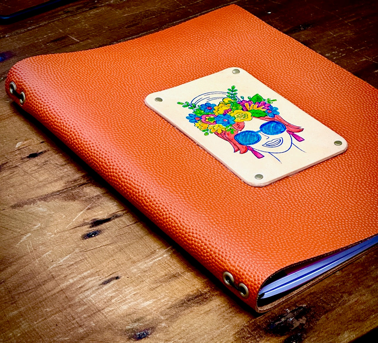low corner angle of beautiful orange pebbled leather journal created by Undercover Popular Leather Goods features a hand painted woman wearing flowers in her hair.